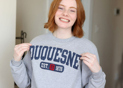 Hannah, a young college student, smiling and holding out her Duquesne college sweatshirt proudly