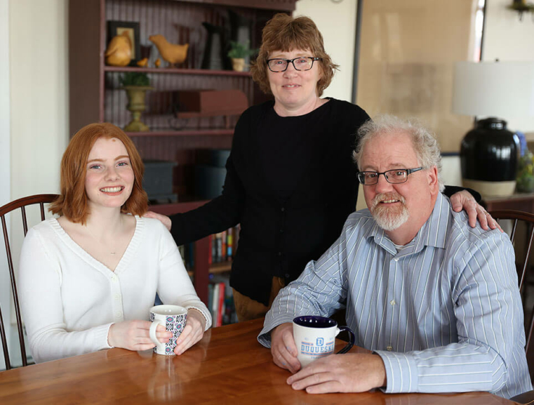 Hannah sitting at her family's kitchen table, while holding a mug with her dad and mom alongside her