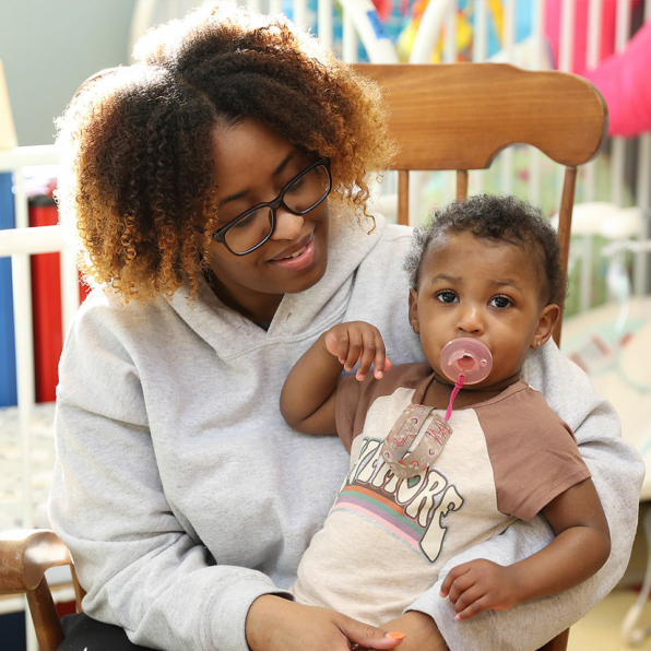 A young woman smiling down on the cutest young toddler sucking on a pacifier