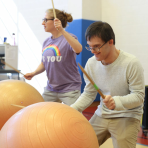 Two adults with special needs joyfully pounding on large exercise balls with drumsticks