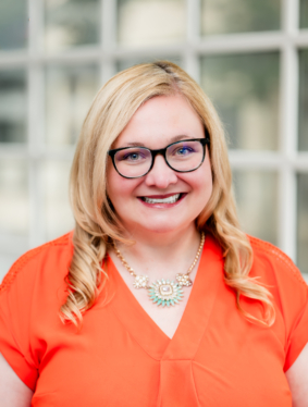 Picture of a woman with Blonde hair wearing glasses in an bright orange shirt.