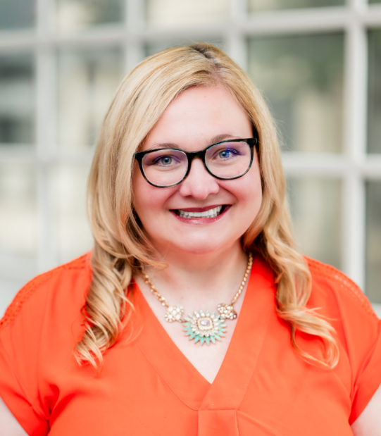 Picture of a woman with Blonde hair wearing glasses in an bright orange shirt.